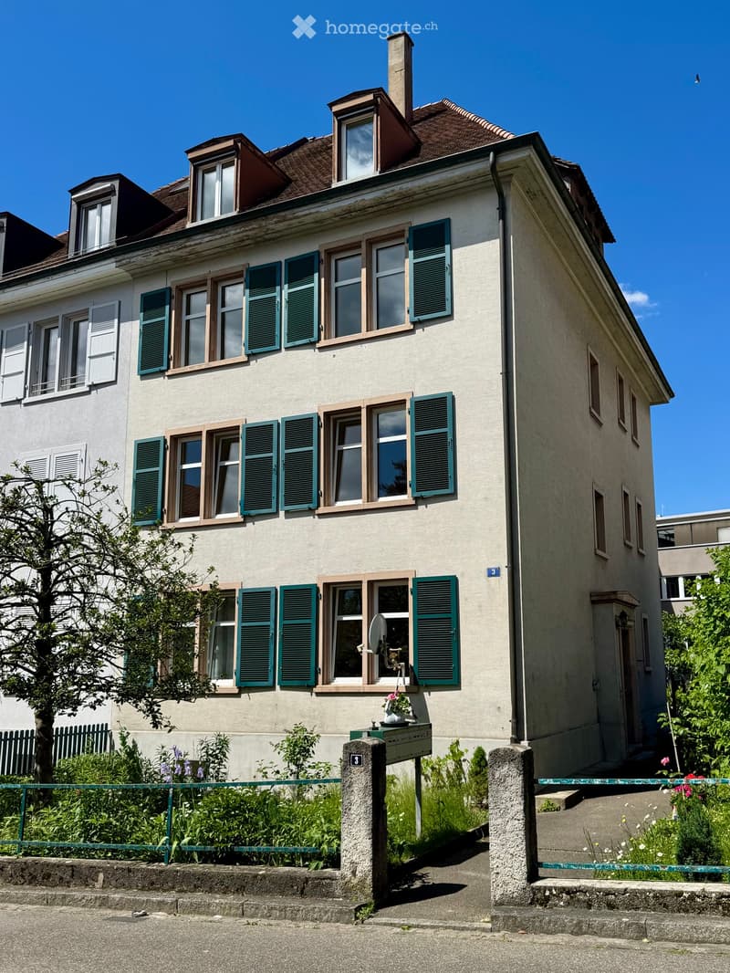 Two-story house with light beige walls, green shutters, multiple windows, and a small garden in front.