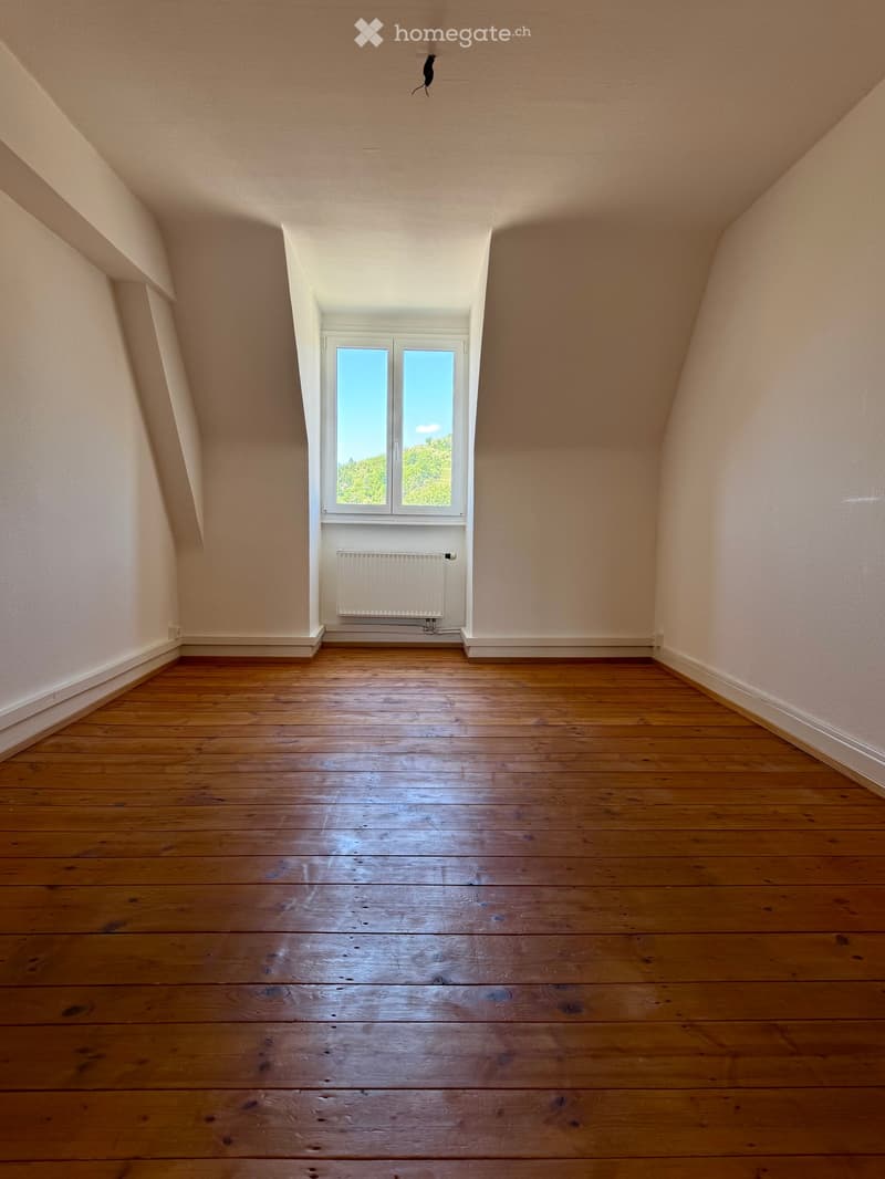 Empty attic room with wooden flooring, white walls, and a single window with a radiator below it.