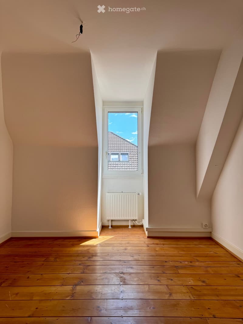 Empty attic room with wooden flooring, slanted ceilings, and a single window showing a view of a roof and sky. A white radiator is mounted on the wall below the window.