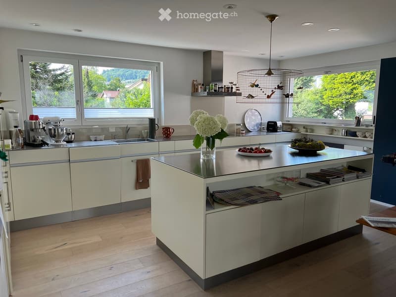 Modern kitchen with light-colored cabinets, a central island, and large windows offering a view of greenery. The island features a vase with white flowers, a bowl of fruit, and a stack of books. The kitchen includes a sink, coffee maker, mixer, and variou