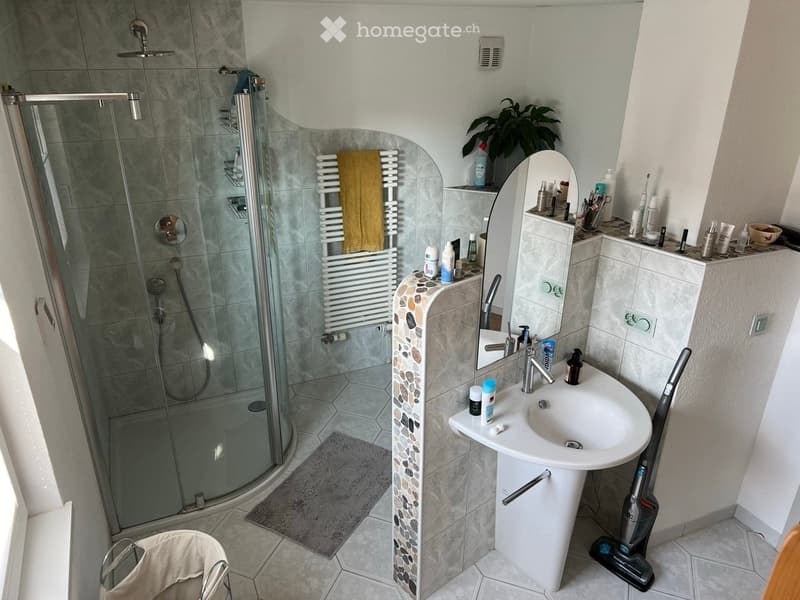 Modern bathroom with a glass-enclosed shower, a white sink with a mirror, a towel rack with a yellow towel, and various toiletries on the counter. A vacuum cleaner is placed on the floor near the sink.
