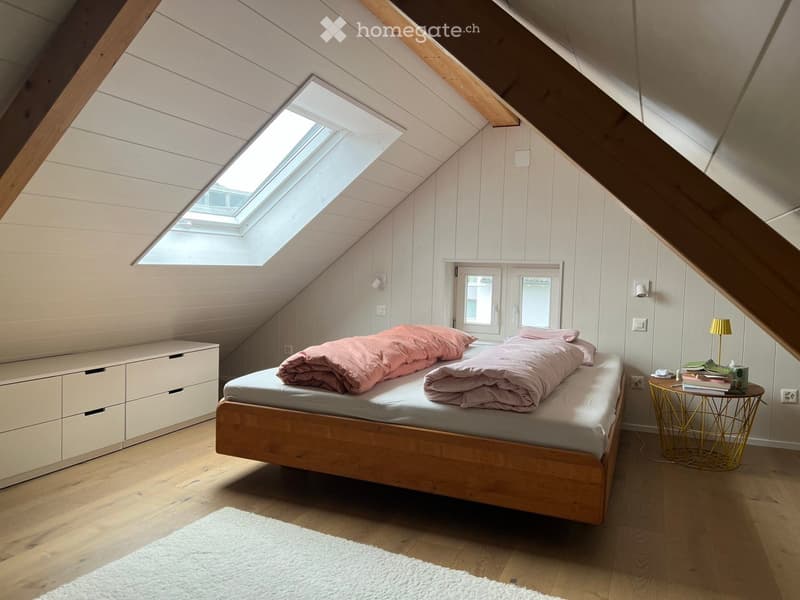 Cozy attic bedroom with wooden floors, slanted wooden ceiling, skylight window, and a bed with pink bedding.