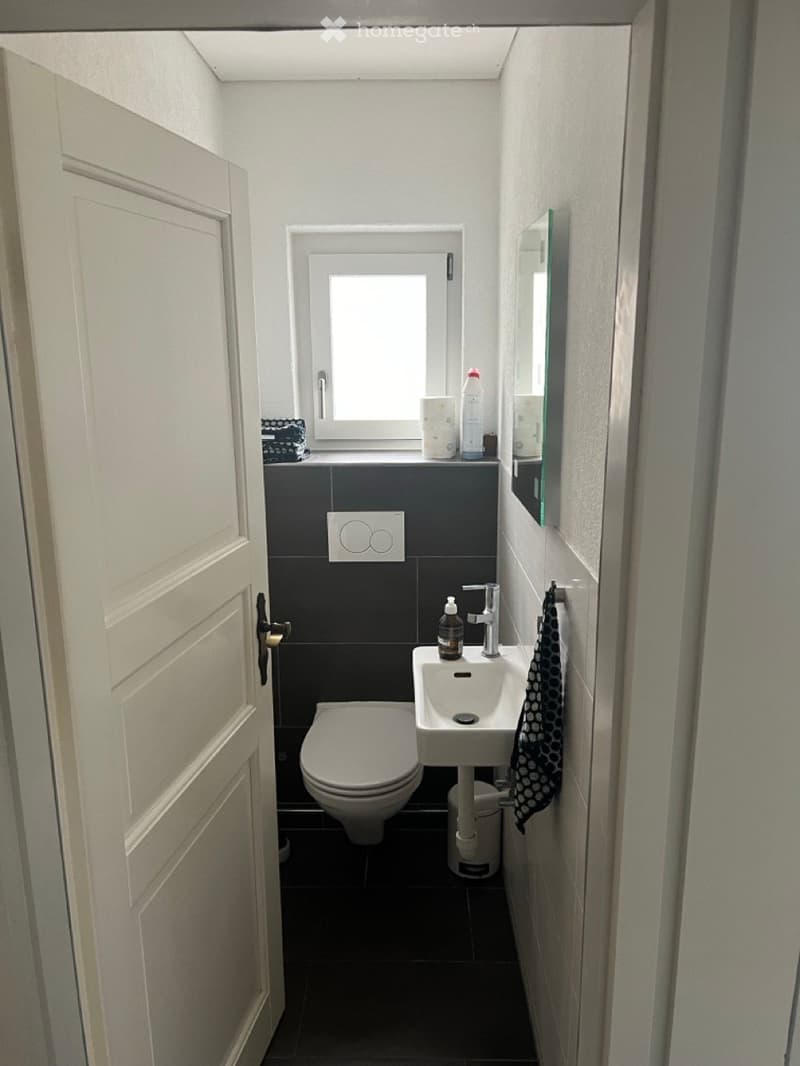 Modern bathroom with dark tiles, white fixtures, a toilet, sink, mirror, and a small window above the sink.