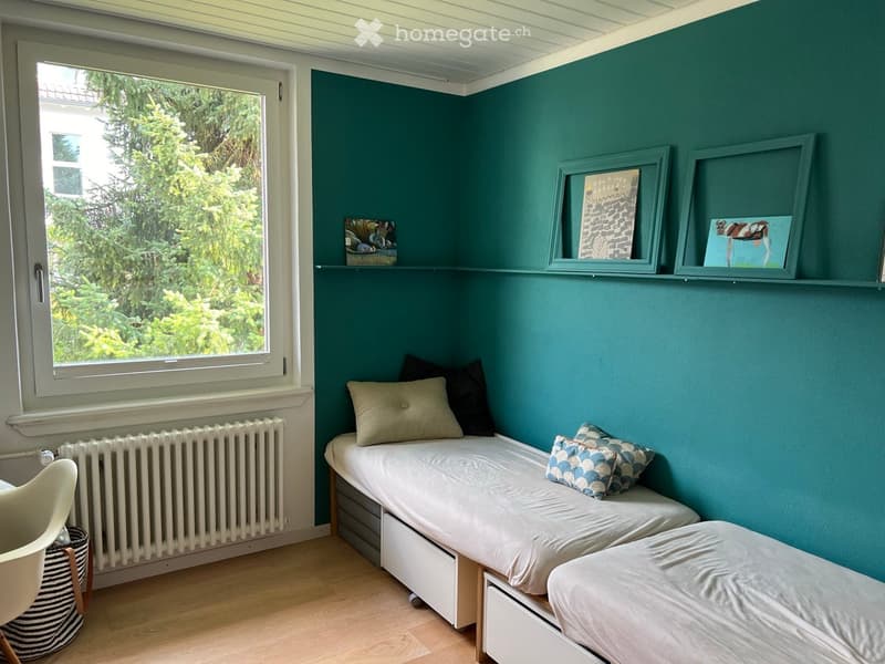 Cozy bedroom with teal walls, two single beds with pillows, a white chair, a radiator, and a shelf with picture frames.