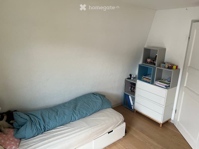 Cozy bedroom with a single bed, nightstand, and shelving unit. The bed is made with blue bedding, and the room features light wooden flooring and white walls.
