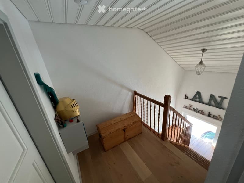 Staircase with wooden steps and handrail, white walls, and a hanging light fixture. A small wooden chest and a decorative helmet are placed on the left side.