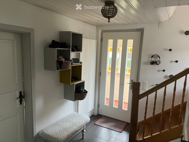 Hallway with a wooden staircase, a white door with glass panels, a small stool, a wall-mounted shelf with baskets and items, and a pair of headphones on the wall.
