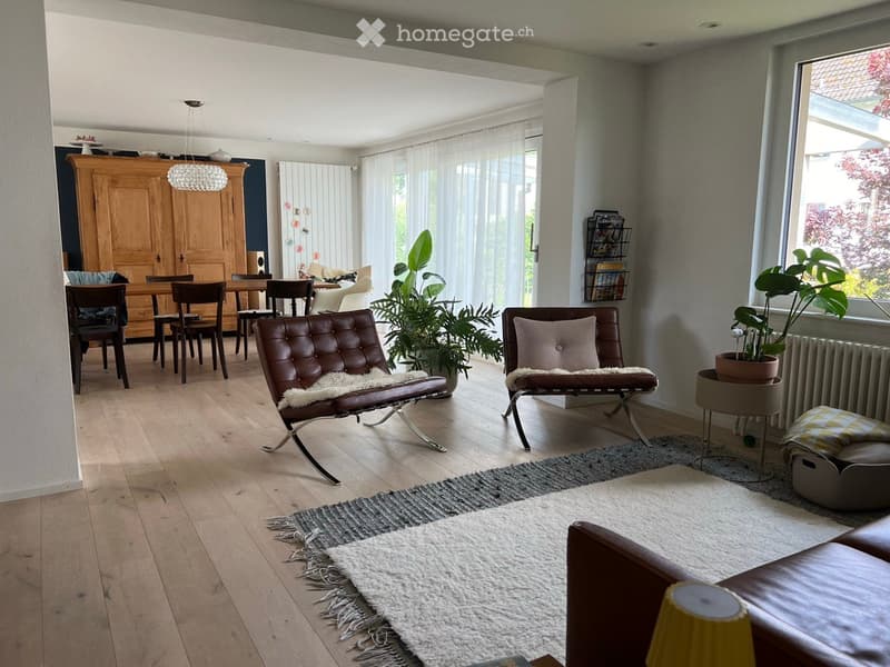Bright living room with wooden flooring, leather sofa, chairs, dining table, and large windows with sheer curtains.