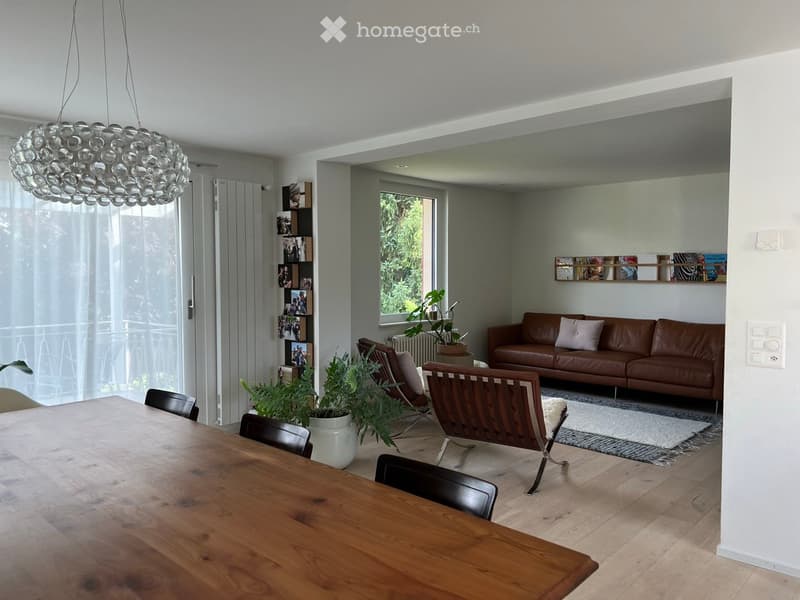 Modern living and dining area with wooden flooring, large windows, a leather sofa, a wooden dining table with chairs, and decorative plants.