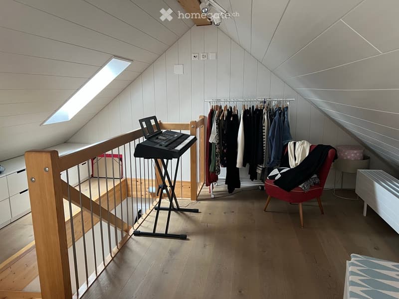 Attic room with wooden floors, slanted ceilings, and large skylight. Features a keyboard, red chair, clothing rack, and laptop on a shelf.