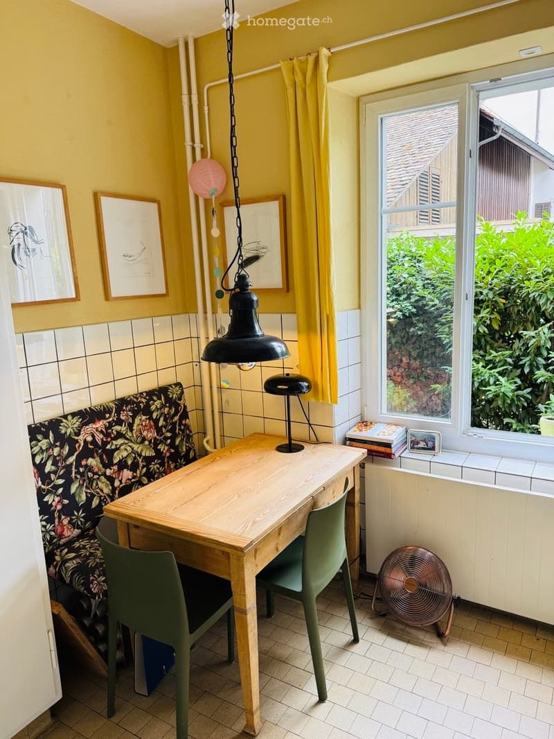 Bright room with yellow walls, wooden table, green chairs, floral-patterned sofa, and large window with yellow curtains.