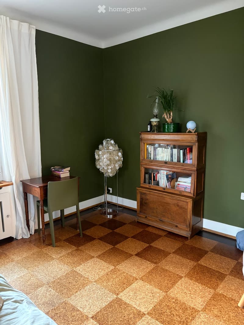 A cozy room with dark green walls, a checkered floor, a wooden bookshelf, a small table with a chair, a floor lamp, and decorative items including a potted plant and a globe.