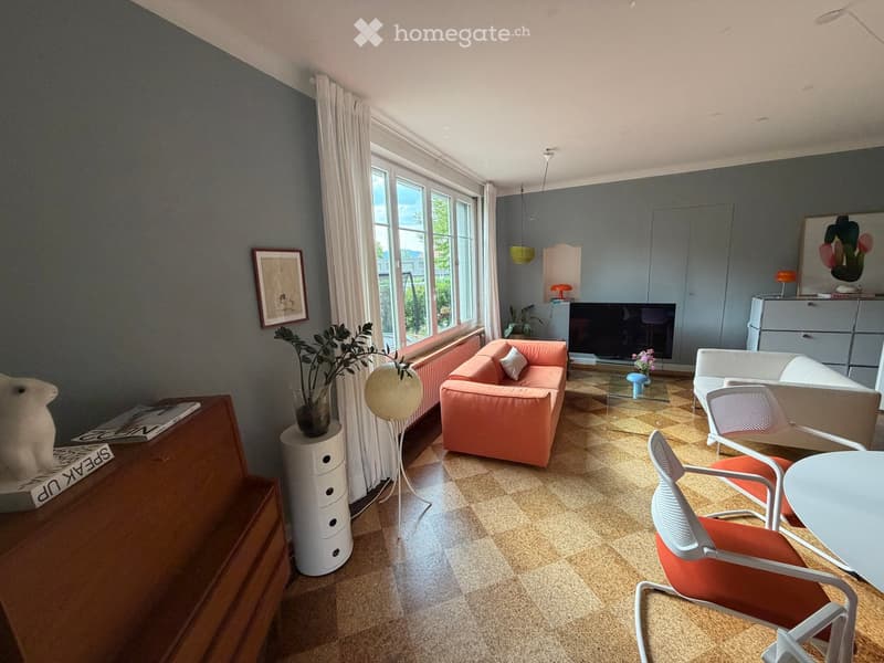 Modern living room with large windows, grey walls, and a mix of contemporary and vintage furniture including a pink sofa, white chairs, a glass coffee table, and a TV stand.