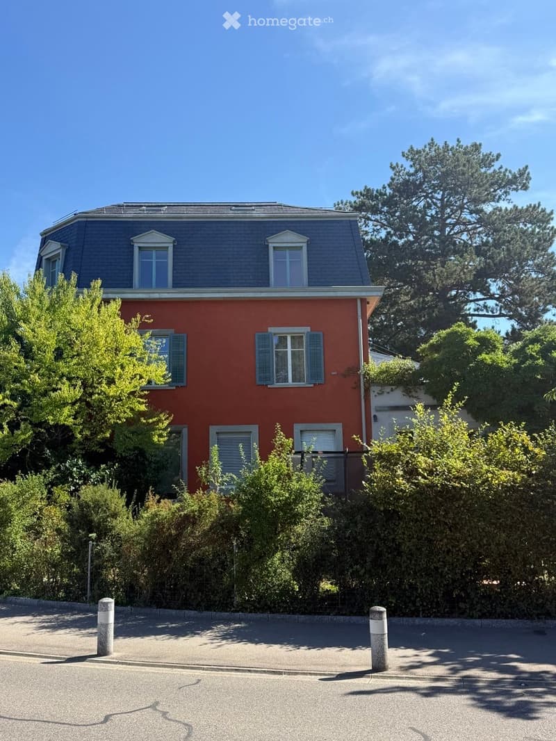 Two-story red house with grey roof, multiple windows, and surrounding greenery.