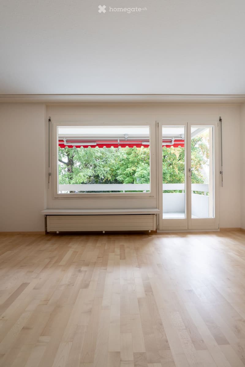 Empty room with light wooden flooring, white walls, large window with red awning, and sliding glass doors leading to a balcony.