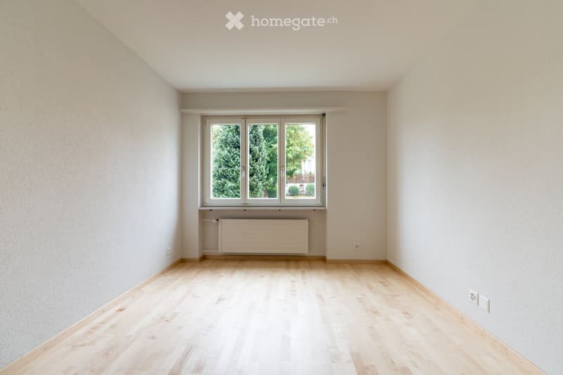 Empty room with light wooden flooring, white walls, and a large window with a radiator below it.