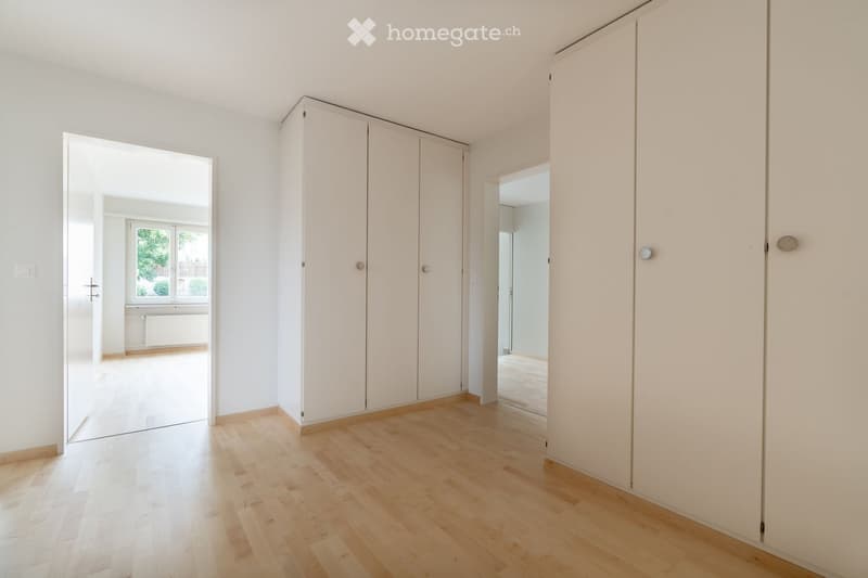 Empty room with light wooden flooring, white walls, built-in wardrobes, and an open doorway leading to another room with a window.