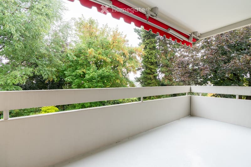 Balcony with white railing, red awning, and lush green trees in the background.