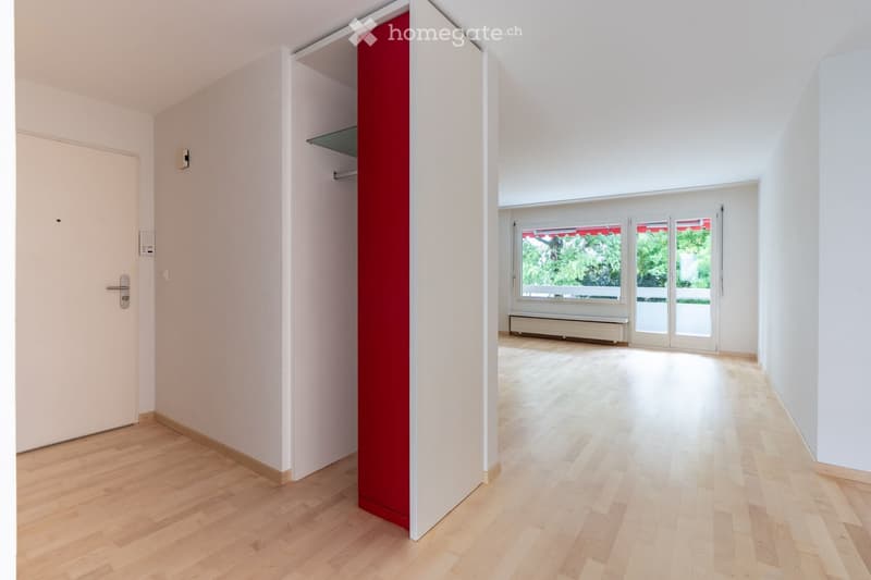Empty room with light wooden flooring, white walls, and a large window with a view of greenery. The room features a sliding glass door leading to a balcony area.