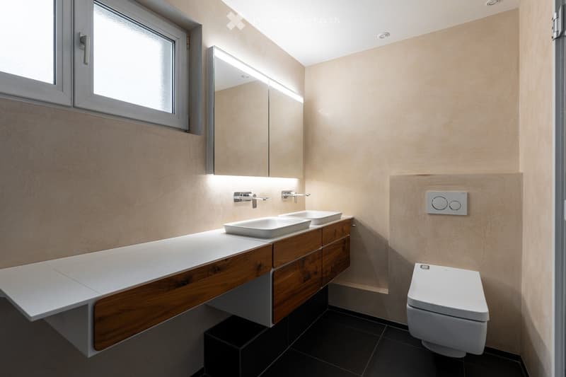 Modern bathroom with a double sink vanity, wooden cabinet, mirror, and toilet. Light-colored walls and dark tiled floor.