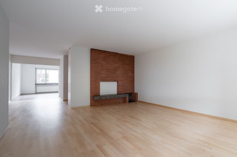 Empty room with light wooden flooring, white walls, and a brick accent wall with a fireplace. The room has large windows and an open doorway leading to another space.