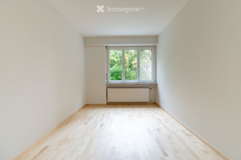 Empty room with light wooden flooring, white walls, and a large window with a radiator below it.