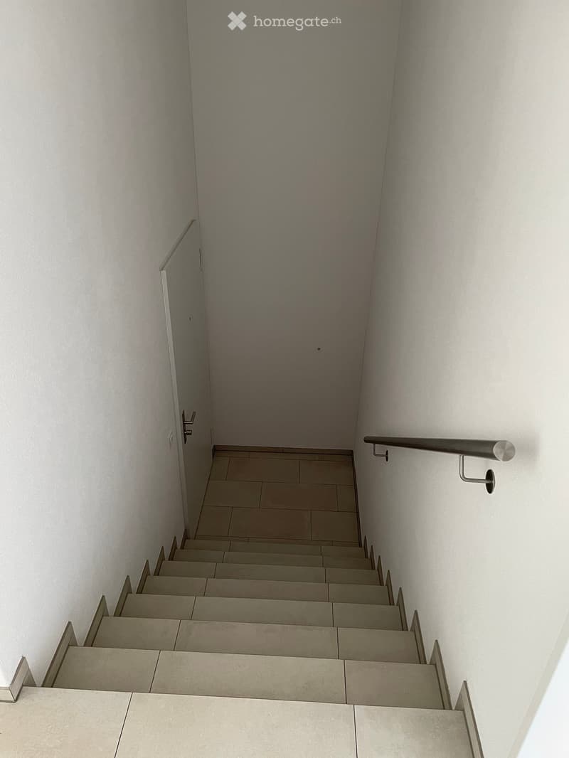 Modern staircase with beige tiled steps and matching walls, featuring a sleek metal handrail on the right side and a glass door at the top.