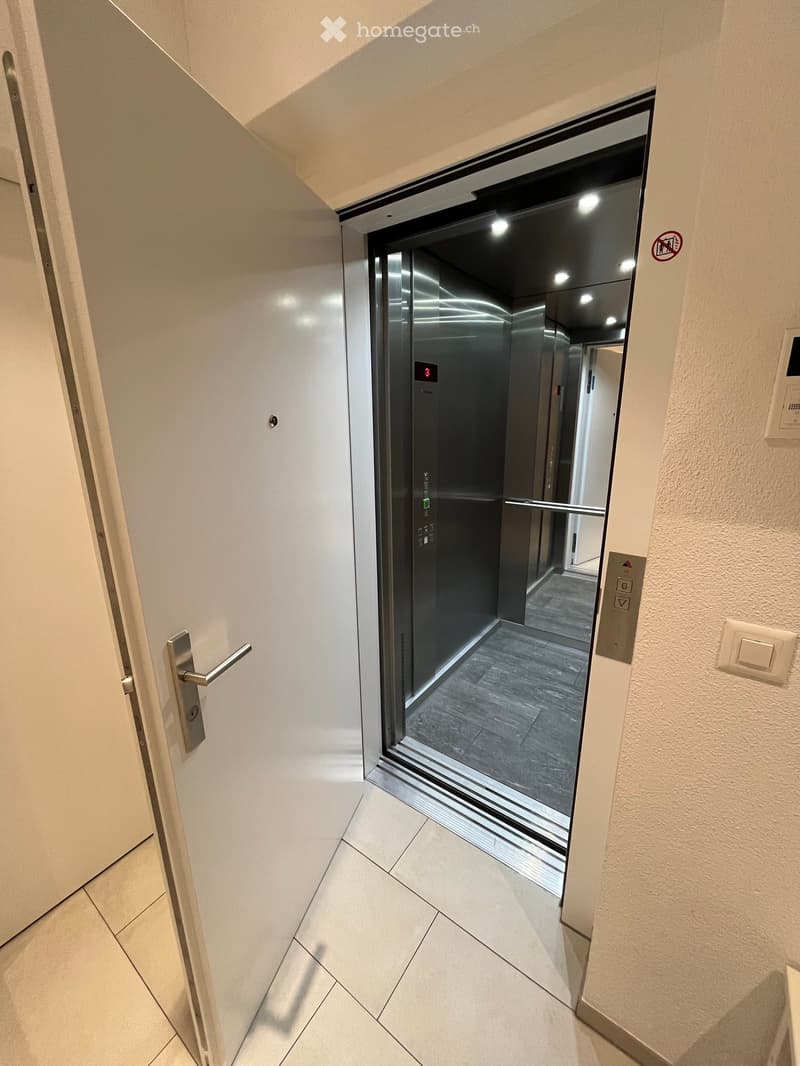 Modern hallway with tiled flooring, elevator door open, showing interior of elevator with gray carpeted floor and control panel.