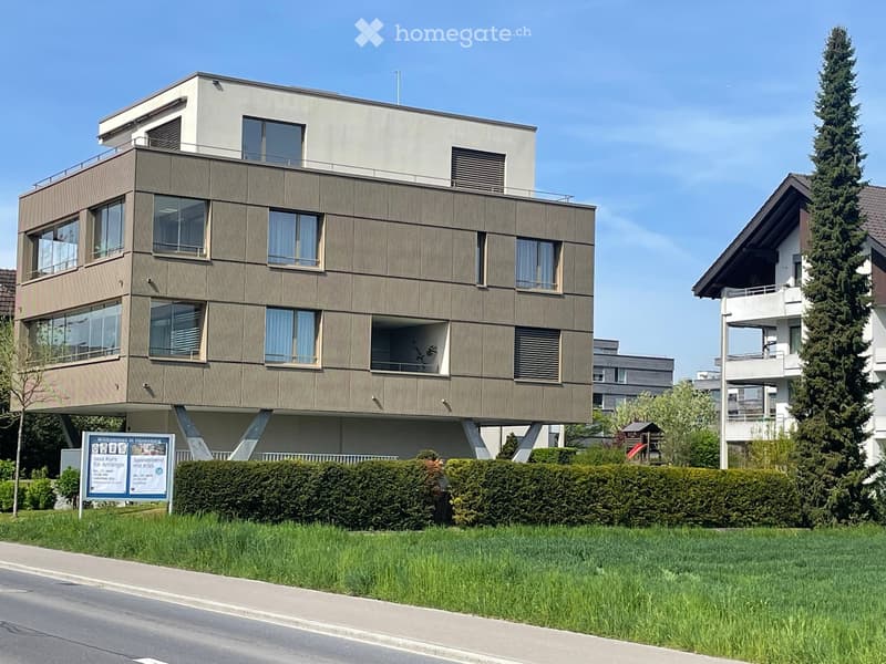 Modern multi-story building with large windows and balconies, surrounded by greenery and a signboard.