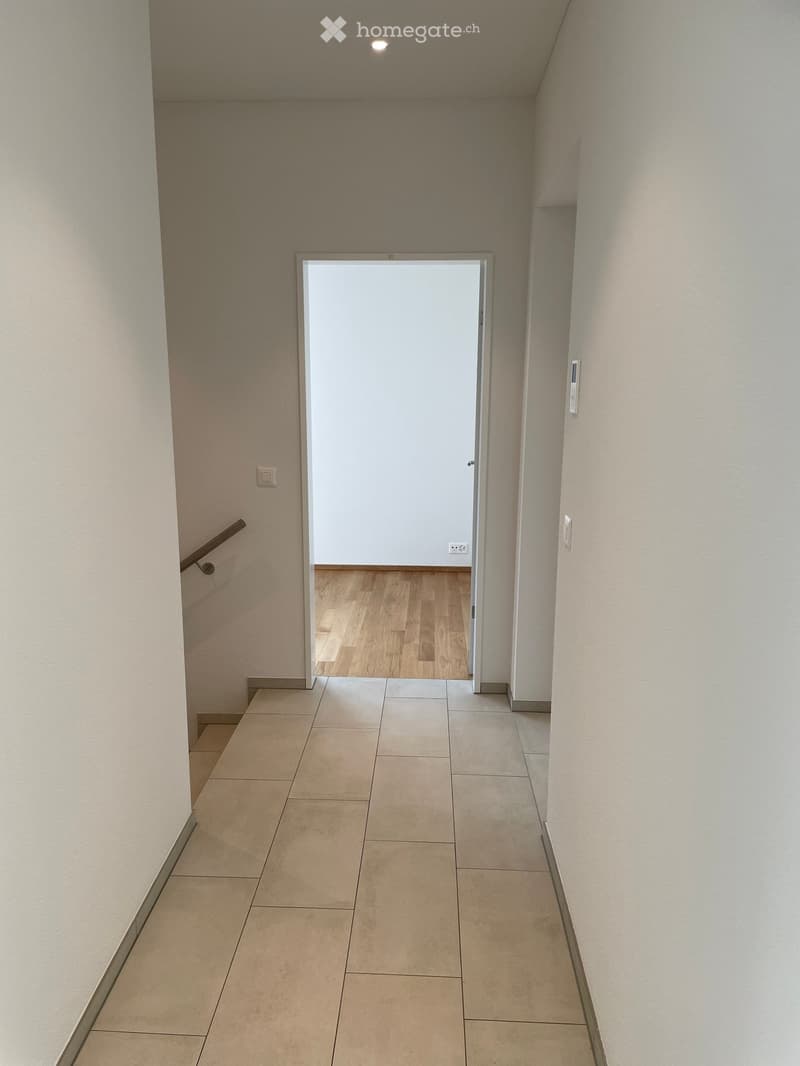 Clean hallway with tiled floor and beige walls, featuring a wooden staircase leading to an open doorway with wooden flooring beyond.