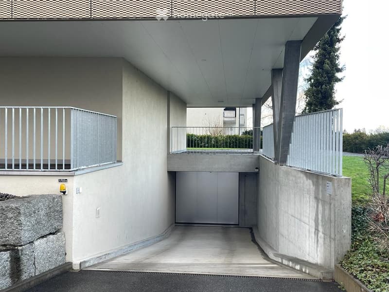 Modern building entrance with a covered garage area, raised ground floor, and balcony with metal railings.
