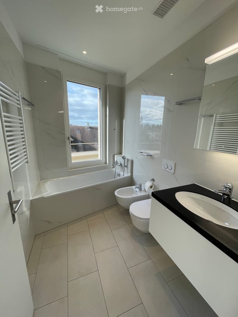 Modern bathroom with a bathtub, toilet, sink, bidet, and radiator. Features large windows with a view of the roof and surrounding area.