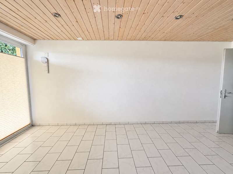 Empty room with white tiled flooring, light-colored walls, and a wooden slatted ceiling. Features a large window with blinds and a door on the right side.