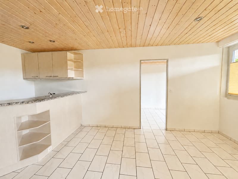 Empty kitchen with white tiled floors, light wooden cabinets, and a granite countertop. The space is bright with natural light from a window and has a doorway leading to another room.