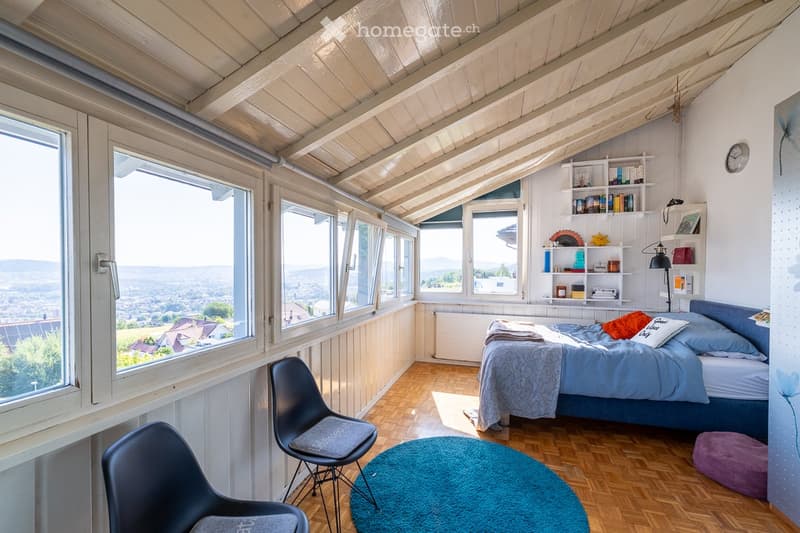 Bright bedroom with wooden slanted ceiling, large windows offering a scenic view, bed with blue bedding, two chairs, round blue rug, and built-in shelving unit.