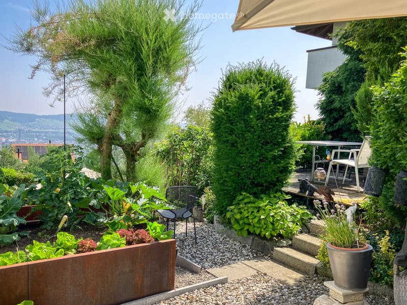 Well-maintained garden with various plants, shrubs, and a small seating area with a table and chairs. The garden features a mix of vegetables, flowers, and decorative elements, with a scenic view of the surrounding landscape.