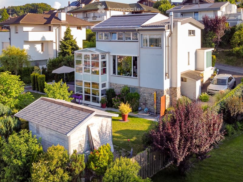 Aerial view of a two-story house with a modern design, surrounded by a well-maintained garden, featuring a greenhouse, patio, and solar panels on the roof.