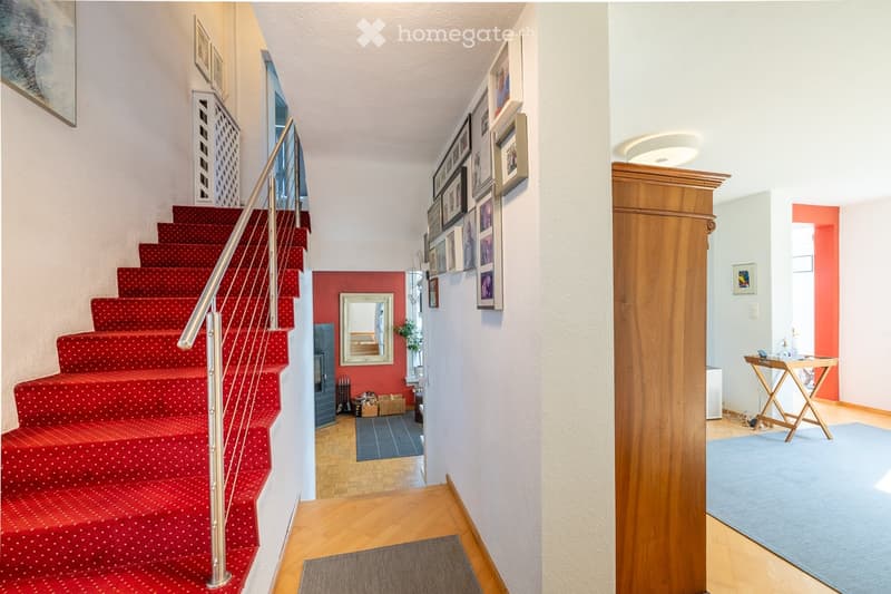 Staircase with red carpeted steps and a metal railing, framed pictures on the wall, a small table with items, and a blue rug in the hallway.