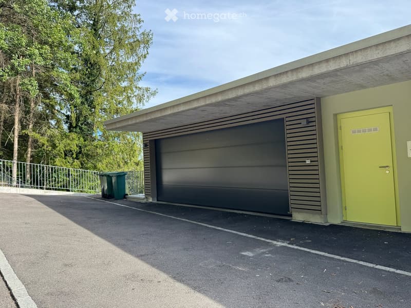 A modern garage with a grey shutter door, a bright green entrance door, and a covered roof area. Green trash bins are placed nearby.