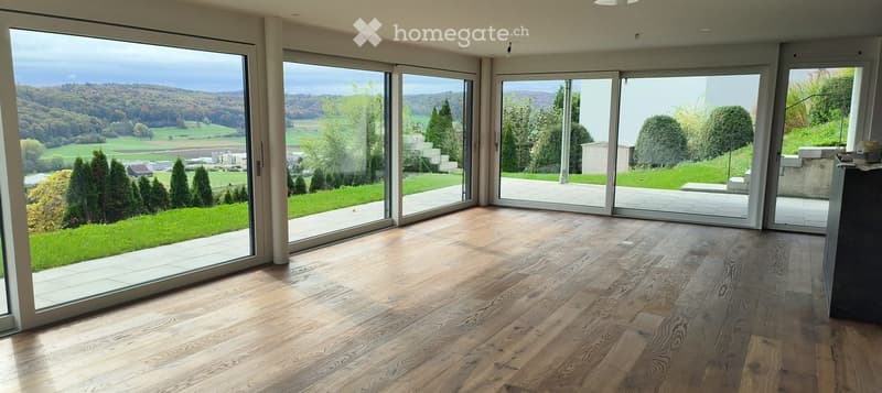 Bright, unfurnished room with large sliding glass doors opening to a landscaped garden, wooden flooring, and expansive scenic views.