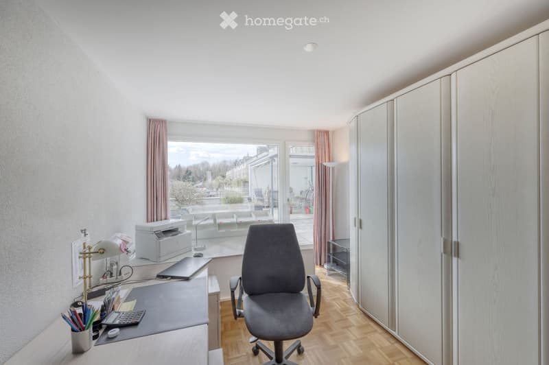 A well-lit office space with a wooden floor, large windows, a desk with a lamp, printer, laptop, and a comfortable office chair. There is a built-in wardrobe and a view of a patio area.