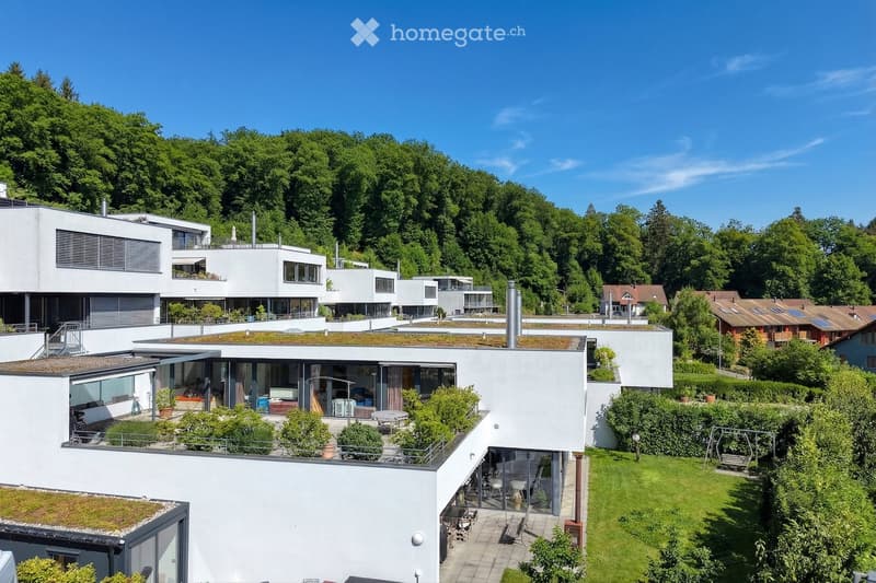 A modern residential complex with multiple white buildings, green roofs, and surrounding greenery. Features include balconies, terraces, and landscaped gardens.