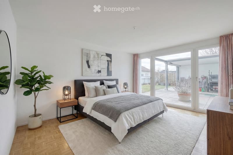 Modern bedroom with a large bed, nightstand, lamp, and a large mirror on the wall. The room features wooden flooring, a white rug, and large windows with pink curtains leading to a patio.