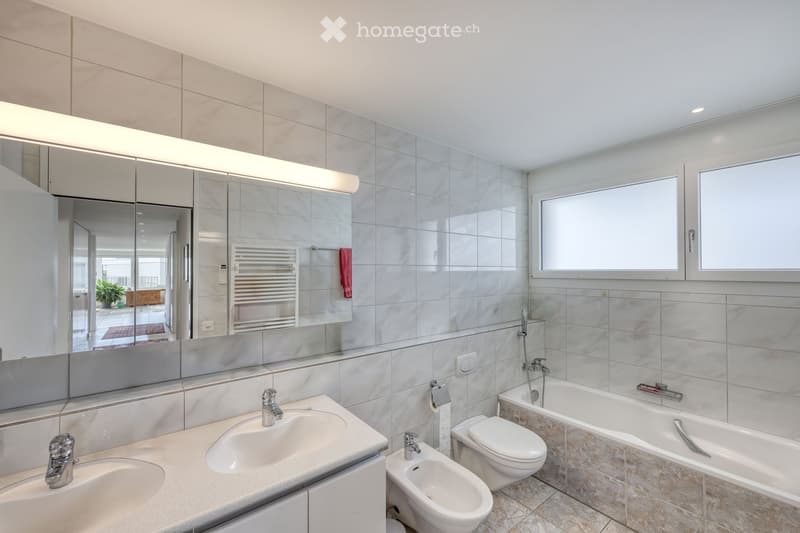 Modern bathroom with double sink, bathtub, toilet, bidet, large mirror, and radiator. Light grey tiles and warm lighting.