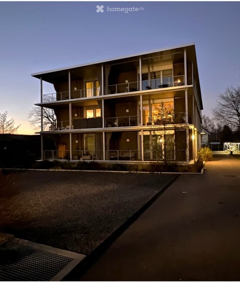 Modern two-story building with large glass windows and balconies, illuminated at dusk, surrounded by a paved area and sparse vegetation.