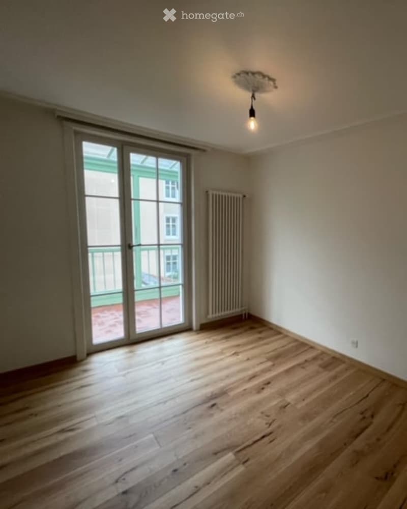 Empty room with wooden flooring, large glass sliding door leading to a balcony, radiator, and a single ceiling light fixture.