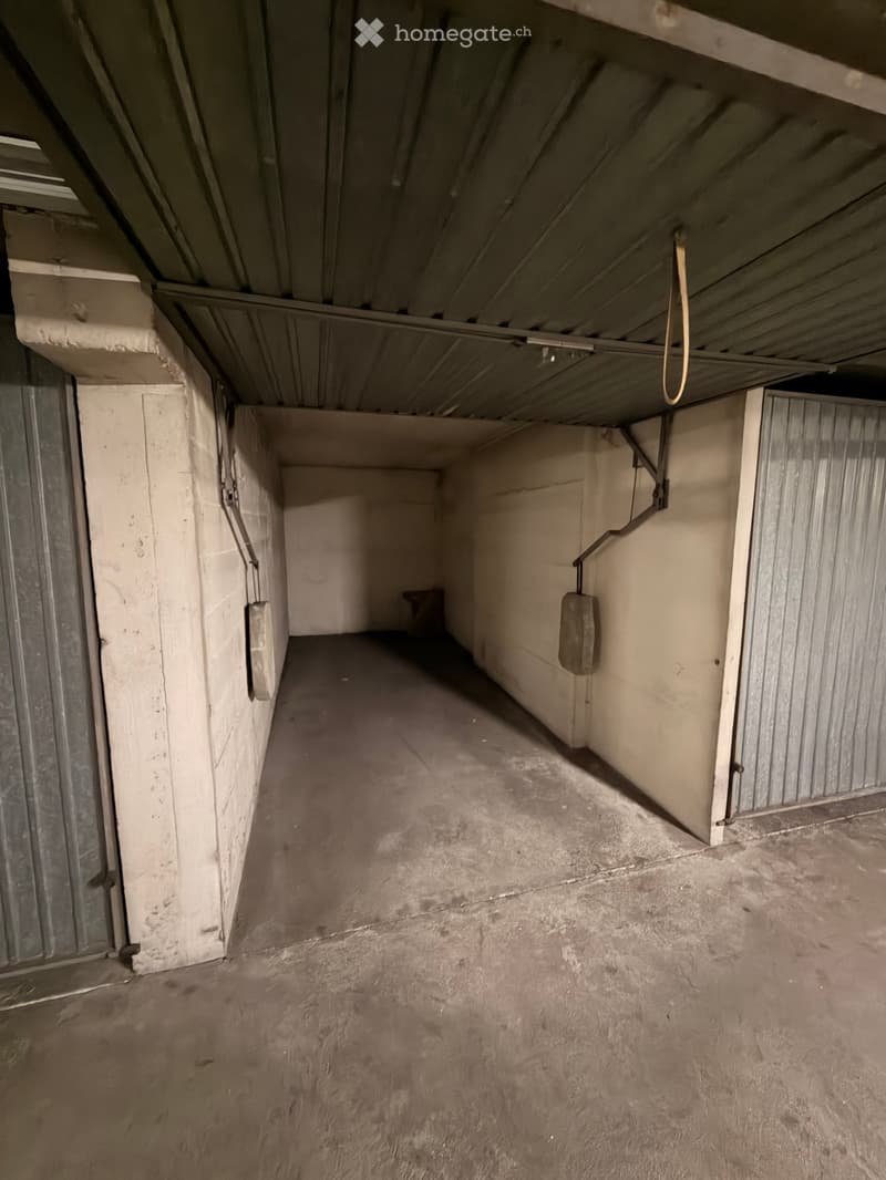 Empty garage with concrete floor, metal walls, and a metal door at the far end. Exposed pipes and a hanging cable are visible.
