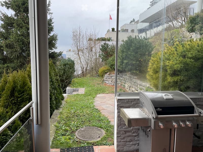 Outdoor patio with a grill, stone walls, manicured shrubs, and a view of the city and water.