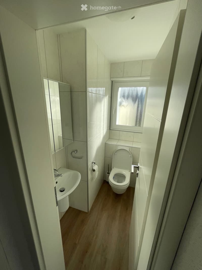 Modern bathroom with white tiles, wooden floor, toilet, sink, and mirror. Features a small window with frosted glass.