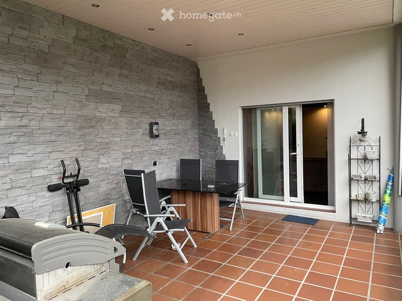 Balcony with tiled floor, stone wall, small table with chairs, exercise bike, and a shelf with decorative items.